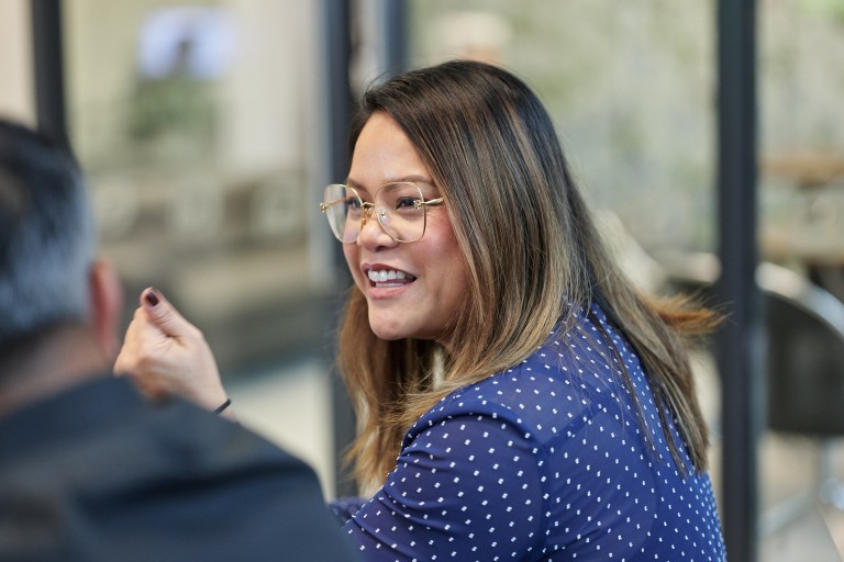 Eine Person sitzt in einem Innenraum und spricht mit einer anderen Person im Vordergrund. Die abgebildete Person trägt ein Oberteil mit Muster und eine Brille. Der Hintergrund zeigt einen unscharfen Bürobereich mit Glaswänden.