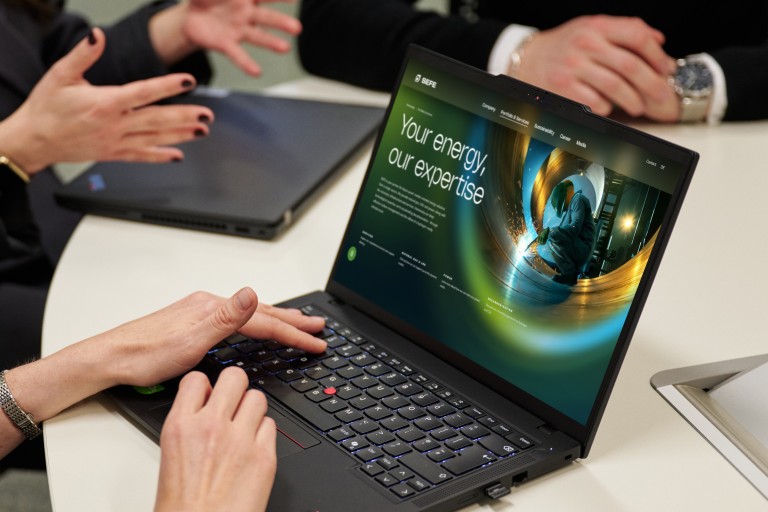 mehrere Personen sitzen an einem runden Tisch in einem Besprechungsraum und arbeiten mit Laptops. Auf einem geöffneten Laptop ist eine Website mit dem Schriftzug ‚Your energy. Our expertise.‘ zu sehen. Hände und Gesten deuten auf eine gemeinsame Diskussion hin
