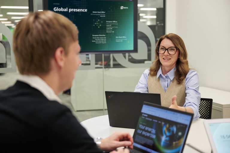Zwei Personen sitzen in einem Besprechungsraum an einem Tisch und arbeiten jeweils an einem Laptop. Eine der Personen zeigt mit einer Handgeste auf die andere. Im Hintergrund ist ein Bildschirm mit einer Darstellung globaler Standorte und Text zur internationalen Präsenz zu sehen.