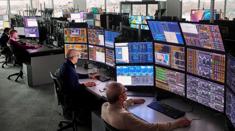 Trading Floor mit mehreren Monitoren, auf denen Marktanalysen und Kursdaten angezeigt werden, Mitarbeitende beobachten Energiedaten mit Blick auf die Skyline einer Großstadt.