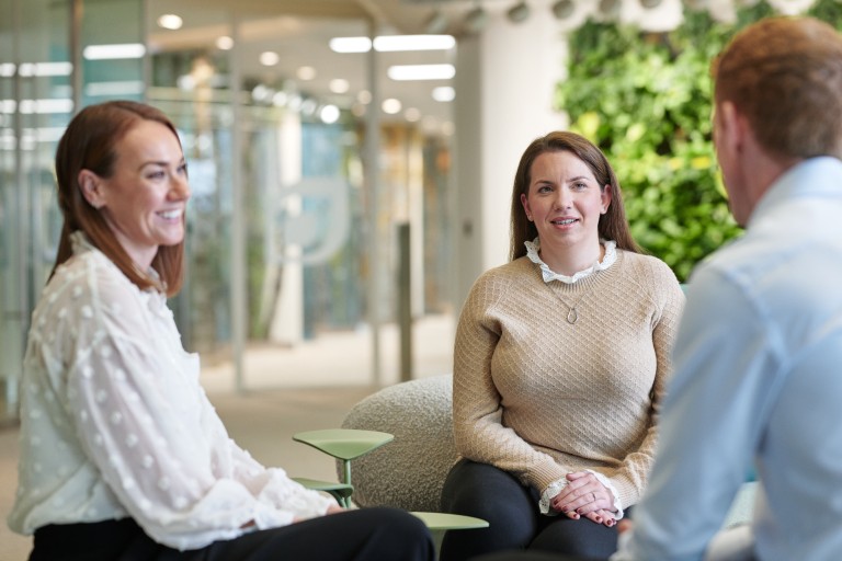 Drei Personen sitzen in einem offenen Bürobereich zusammen. Zwei Personen sitzen nebeneinander auf Sesseln und blicken zu einer dritten Person, die ihnen gegenübersitzt. Im Hintergrund sind Glaswände, Pflanzen und ein modern gestalteter Innenraum zu sehen.